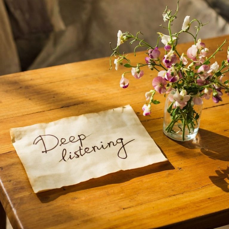 Deep listening. Honey coloured wooden table, small jar of garden of flowers, piece of parchment that says 'Deep Listening' in pen and ink.